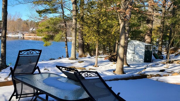 A lakeside deck with two chairs and a table, surrounded by snow and trees.