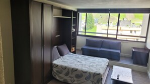 A compact room with a bed, a sofa, a small table, and a large window offering a view of a green landscape.