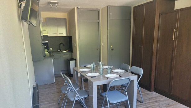 A dining area with a table set for four, four foldable chairs, a kitchen in the background, and a wardrobe with a mirror.