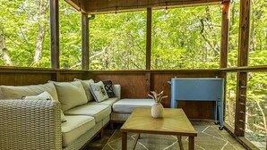 A cozy room with a wicker sofa, a small table, and a view of lush greenery through large windows.