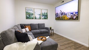 Ein Wohnzimmer mit einem grauen Ecksofa, einem flachbildfernseher an der Wand und drei gerahmten Landschaftsbildern über der Couch.