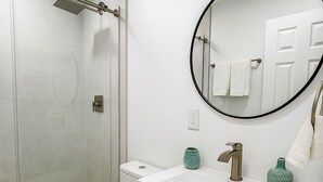 A modern bathroom with a glass-enclosed shower, a white toilet, a white sink with a round mirror, and a cabinet with two doors.
