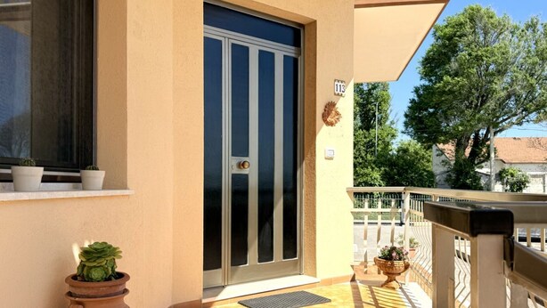 A building with a beige exterior, a sliding glass door, a small porch with a metal railing, and a potted plant on the ground. - PP06 - Porto Potenza, comfortable three-room apartment a few steps from the sea (Porto Potenza Picena)