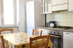 A kitchen with a table covered by a yellow tablecloth, wooden chairs, a refrigerator, a stove, and cabinets. - P061 – Porto Recanati, spacious and comfortable four-room apartment (Porto Recanati)
