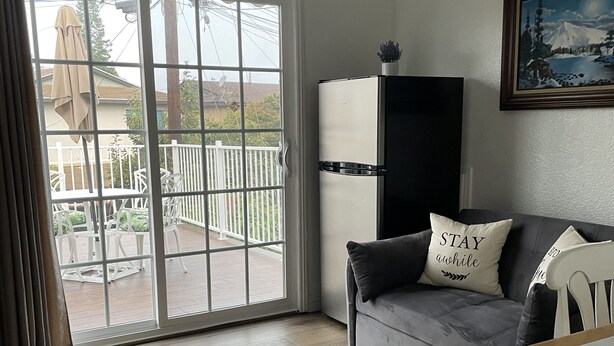 Una sala moderna con un sofá gris, una silla blanca, una mesa de comedor de madera, un refrigerador blanco y negro, y un ventilador de techo.