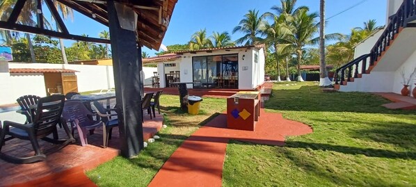 A covered outdoor area with a dining table and chairs, a small building with a tiled roof, and a staircase leading to a second floor.