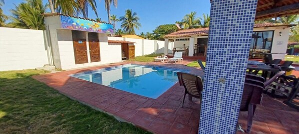 A swimming pool with a tiled deck, a covered patio area, and outdoor seating.