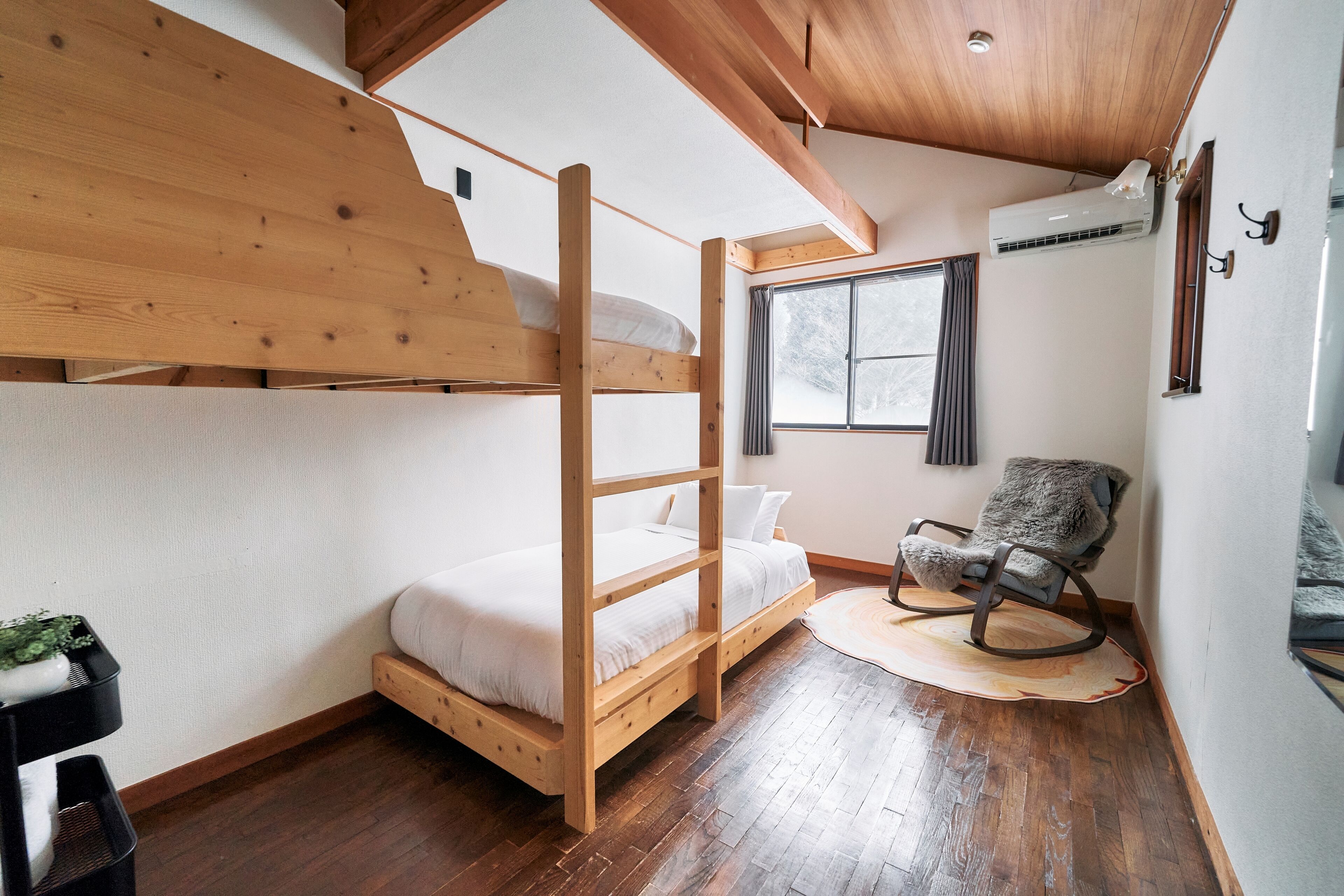 A bunk bed with a wooden frame, a single bed, a rocking chair, a window with curtains, and an air conditioning unit.