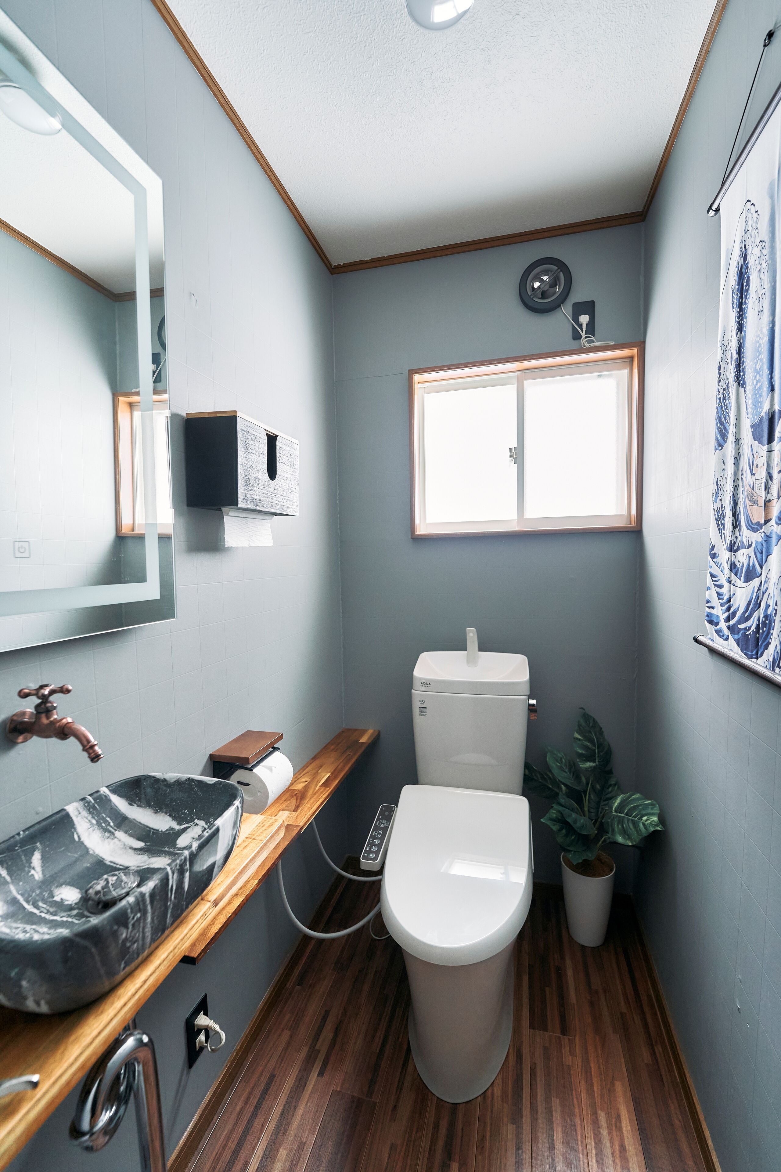 A compact bathroom with a wooden vanity, a white toilet, a wall-mounted sink, a potted plant, and a window.