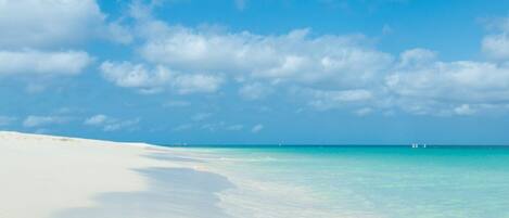 A beach with white sand and clear turquoise water under a blue sky with scattered clouds.