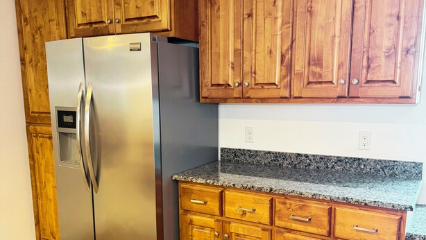 A kitchen with wooden cabinets, a stainless steel refrigerator, granite countertops, and a wooden floor.