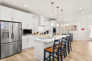 A modern kitchen with white cabinetry, stainless steel appliances, a central island with bar stools, pendant lighting, and a view of the dining area.