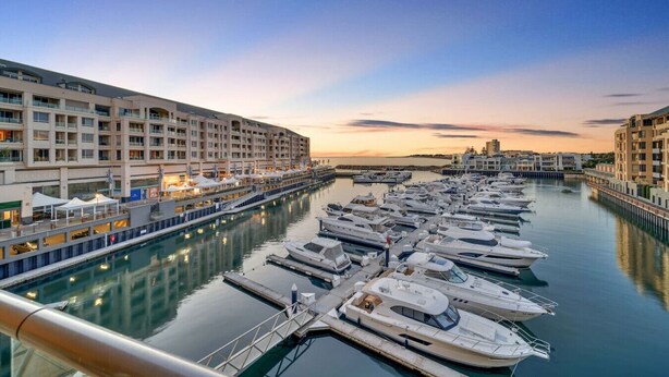 A marina with multiple yachts docked, a waterfront hotel with balconies, and a clear sky.