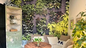 A room with a wall mural of foliage, a green cabinet with shelves, a round wooden table, and a sofa with cushions.