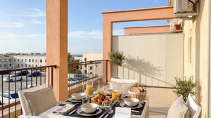Un balcón con una mesa dispuesta para el desayuno, que incluye frutas, pan y jugo, con vistas a un paisaje urbano.