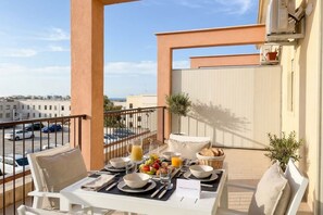 Un balcón con una mesa dispuesta para el desayuno, que incluye frutas, pan y jugo, con vistas a un paisaje urbano.