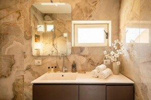 Un baño con pared de mármol, un lavamanos con artículos de aseo, un espejo y un florero con flores.