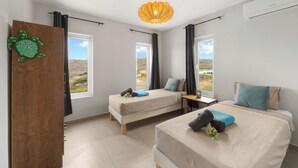 A bedroom with two beds, a wooden door with a welcome sign, a table with a lamp, and large windows with a view of the countryside. - Casa Tortuga – Luxury Holiday Home in Papaya Hill Resort, Cu (Grote Berg)