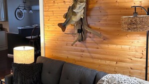 A living room with a dark grey sofa, a lamp, a decorative fish mounted on the wall, and a rustic wooden floor.