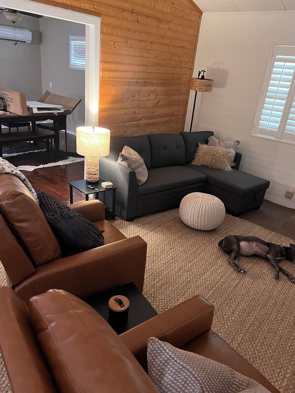 Una sala de estar con un sofá de cuero marrón, un seccional gris oscuro y un otoman redondito. Un perro está acostado en el suelo.