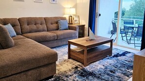 Un salon avec un canapé marron, une table basse en bois et une porte vitrée donnant sur une terrasse extérieure équipée d’une table et de chaises.