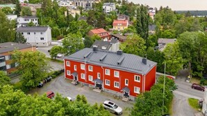 Ein rotes Gebäude mit vielen Fenstern und einer flachen Dachfläche, umgeben von Grünflächen und weiteren Wohnhäusern.