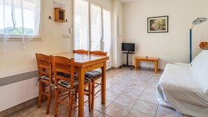 A room with a wooden dining table and chairs, a television, a small wooden side table, a floor lamp, and a bed. - Les Mimosanes by Interhome (Bormes-les-Mimosas)