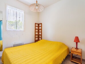 A room with a bed, a window with curtains, a radiator, a wooden side table with a lamp, and a corner shelf.