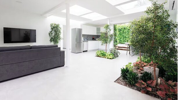 A modern living space with a grey sofa, a flat-screen TV, a kitchen area, and a small garden with plants and a tree. - 3-bedroom 4 Bathrooms Solar Panels, Pool, Near Beaches, Vega Alta / Dorado (Vega Alta)