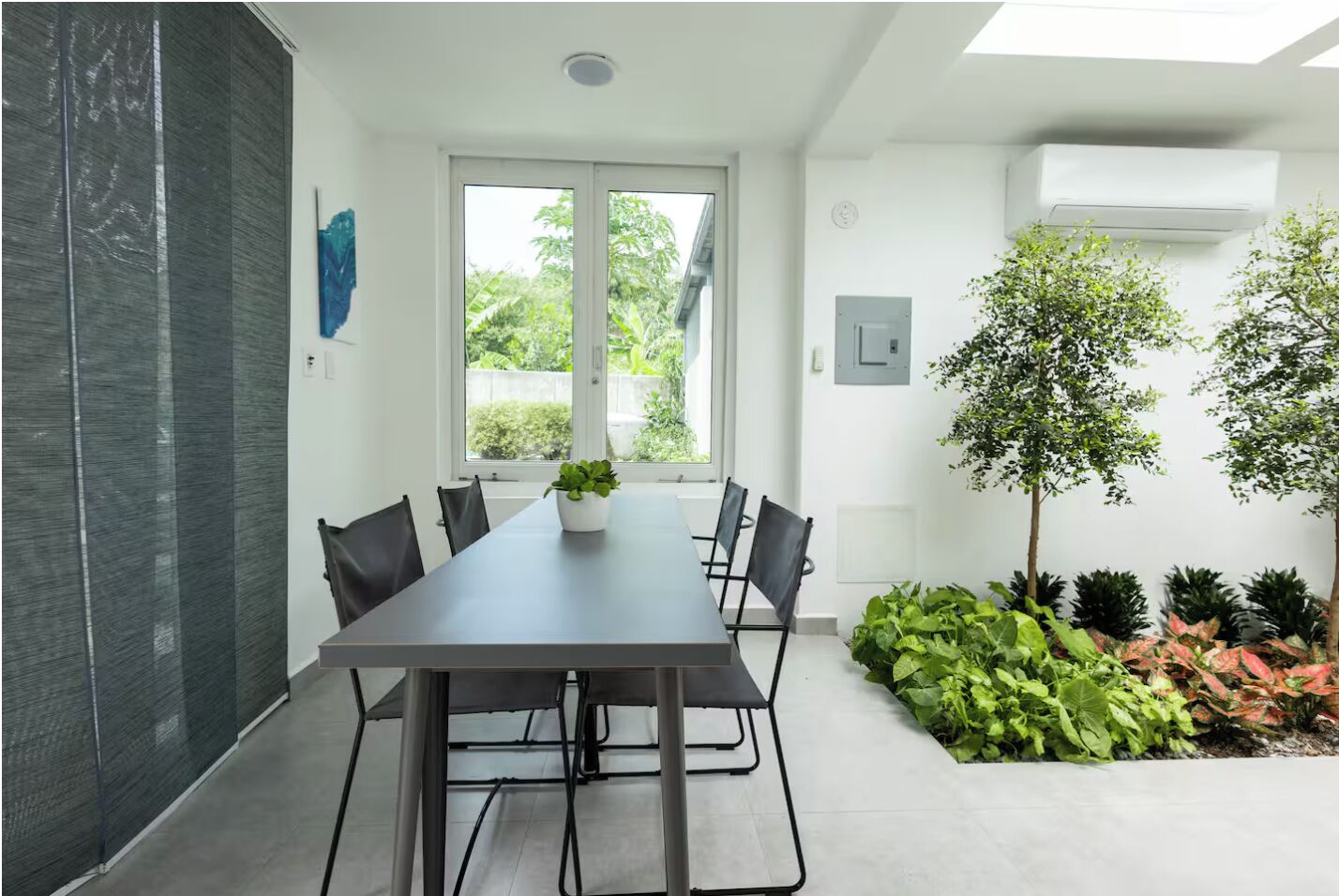 A modern meeting room with a long table, black chairs, a potted plant, and a view of greenery outside.