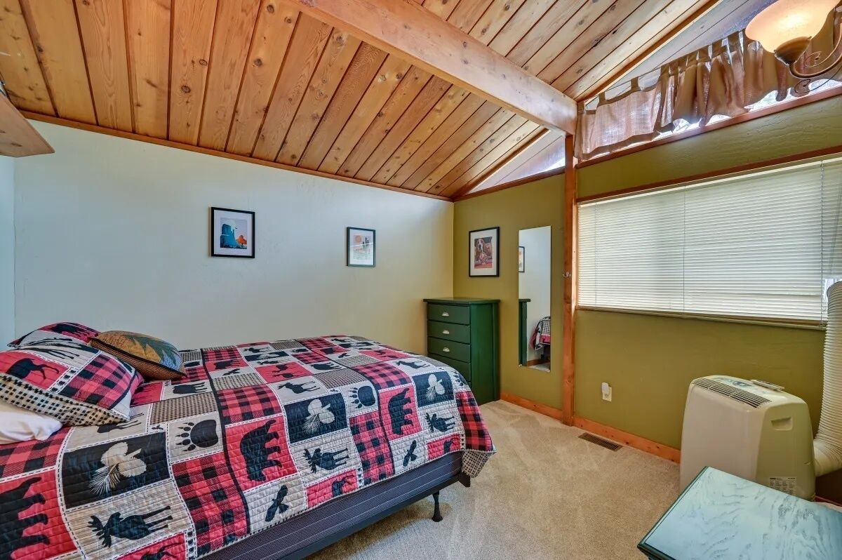 A bedroom with a wooden ceiling, a bed with a quilt, a dresser, and a window with blinds.