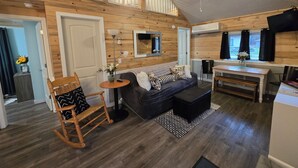 A cozy cabin-style room with a wooden ceiling, a sofa, a rocking chair, a small table with a lamp, and a dining area with a table and chairs.