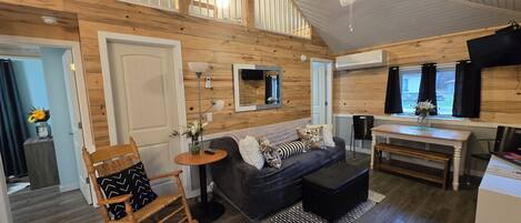A cozy cabin-style room with a wooden ceiling, a sofa, a rocking chair, a small table with a lamp, and a dining area with a table and chairs.