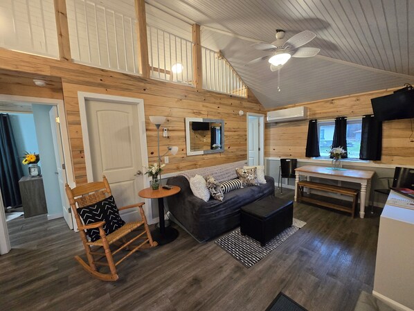 A cozy cabin-style room with a wooden ceiling, a sofa, a rocking chair, a small table with a lamp, and a dining area with a table and chairs.
