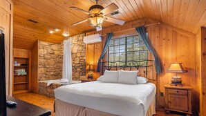 A cozy wooden cabin bedroom with a stone wall, a large bed, a stone fireplace, and a ceiling fan.