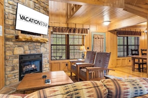 Interior acogedor de una cabaña con chimenea de piedra, paredes de madera, un televisor con la etiqueta 'VACATION STAR' y un comedor.