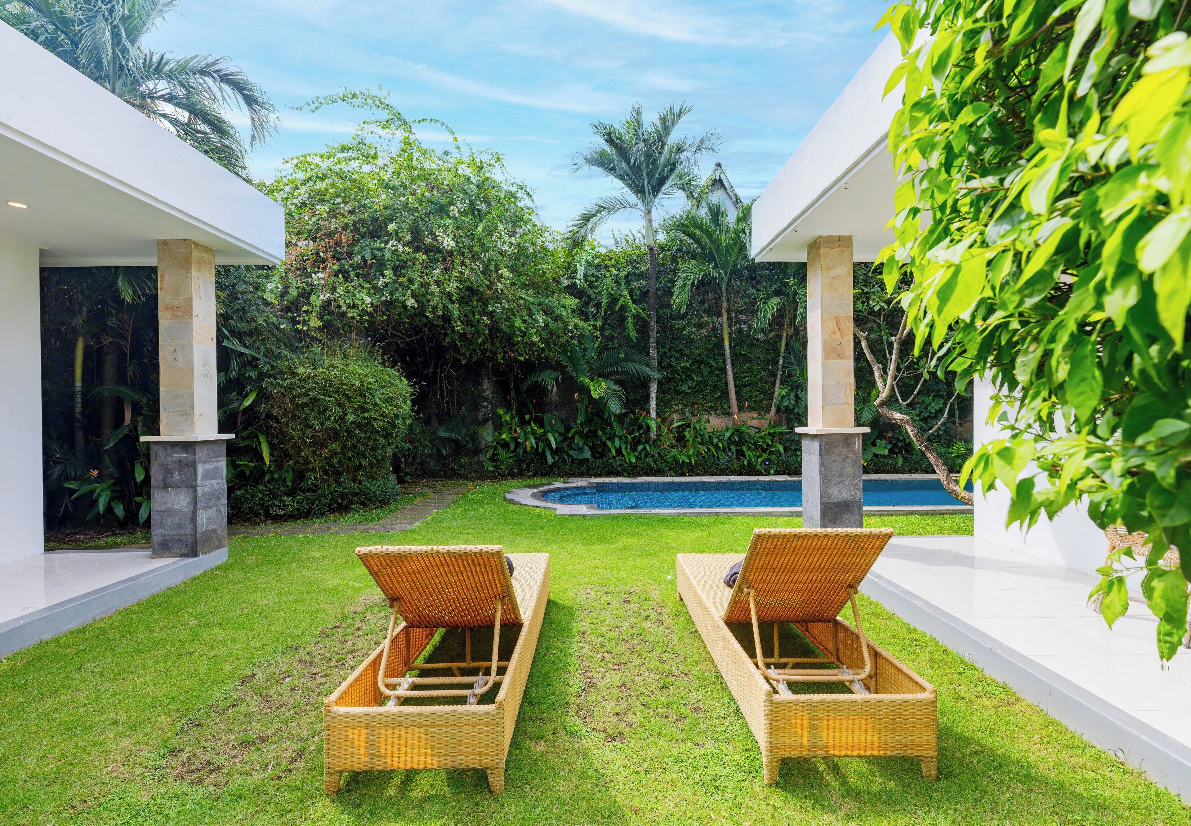 A modern outdoor area with two wicker lounge chairs facing a pool, surrounded by lush greenery and white walls.