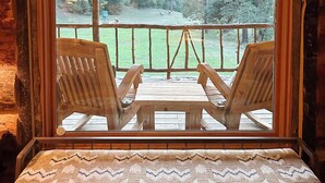 A wooden bench with a woven cushion, a wooden table, and two wooden chairs facing a glass window overlooking a grassy area with trees and a wooden deck.
