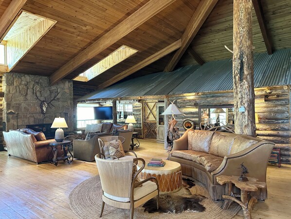 A spacious log cabin interior with a high ceiling, wooden beams, a stone fireplace, leather furniture, and rustic decor. - Skyline Ranch (Cloudcroft)