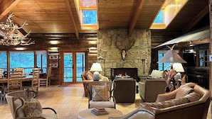 A spacious living room with wooden beams, stone walls, and a stone fireplace. There are several leather sofas and chairs, a wooden coffee table, and a rug on the floor.