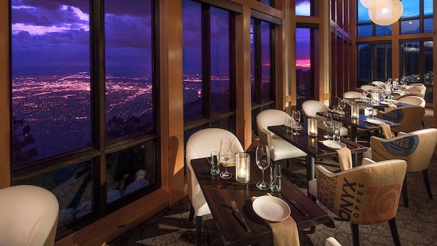 Un restaurant offrant une vue sur le paysage urbain au crépuscule. Les tables sont dressées avec des verres à vin et des assiettes.