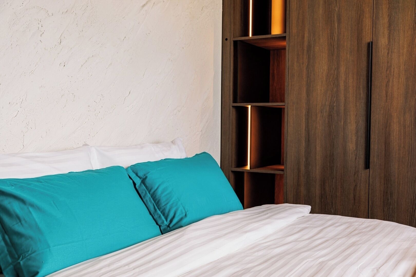 A bed with white and teal bedding, a wooden wardrobe with a vertical handle, and a textured wall.