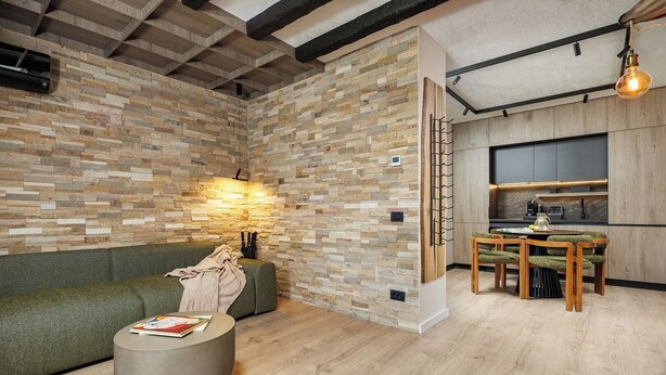 Een moderne woonruimte met een stenen muur, een groene bank, een ronde salontafel en een eethoek met een houten tafel en stoelen.