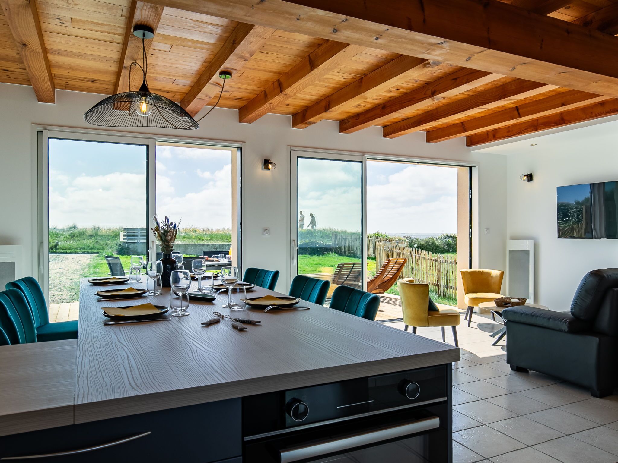 Eine moderne Küche mit einer großen Kochinsel, Holzbalkendecke und großen Fenstern, die Garten und Himmel zeigen.