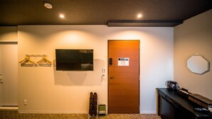 Habitación de hotel con puerta de madera, televisor de pantalla plana en la pared, perchero con ganchos, zapatero, escritorio con cafetera y un pequeño cubo de basura.