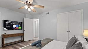 A hotel room with a bed, a TV mounted on the wall, a ceiling fan, and a wooden console table. - Stylish & Comfy | King Bed | Near Love Field | DT (Dallas)