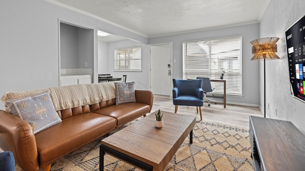 A modern living room with a brown leather sofa, a wooden coffee table, and a blue armchair. There is a TV mounted on the wall, a floor lamp, and a patterned rug. - Stylish & Comfy | King Bed | Near Love Field | DT (Dallas)