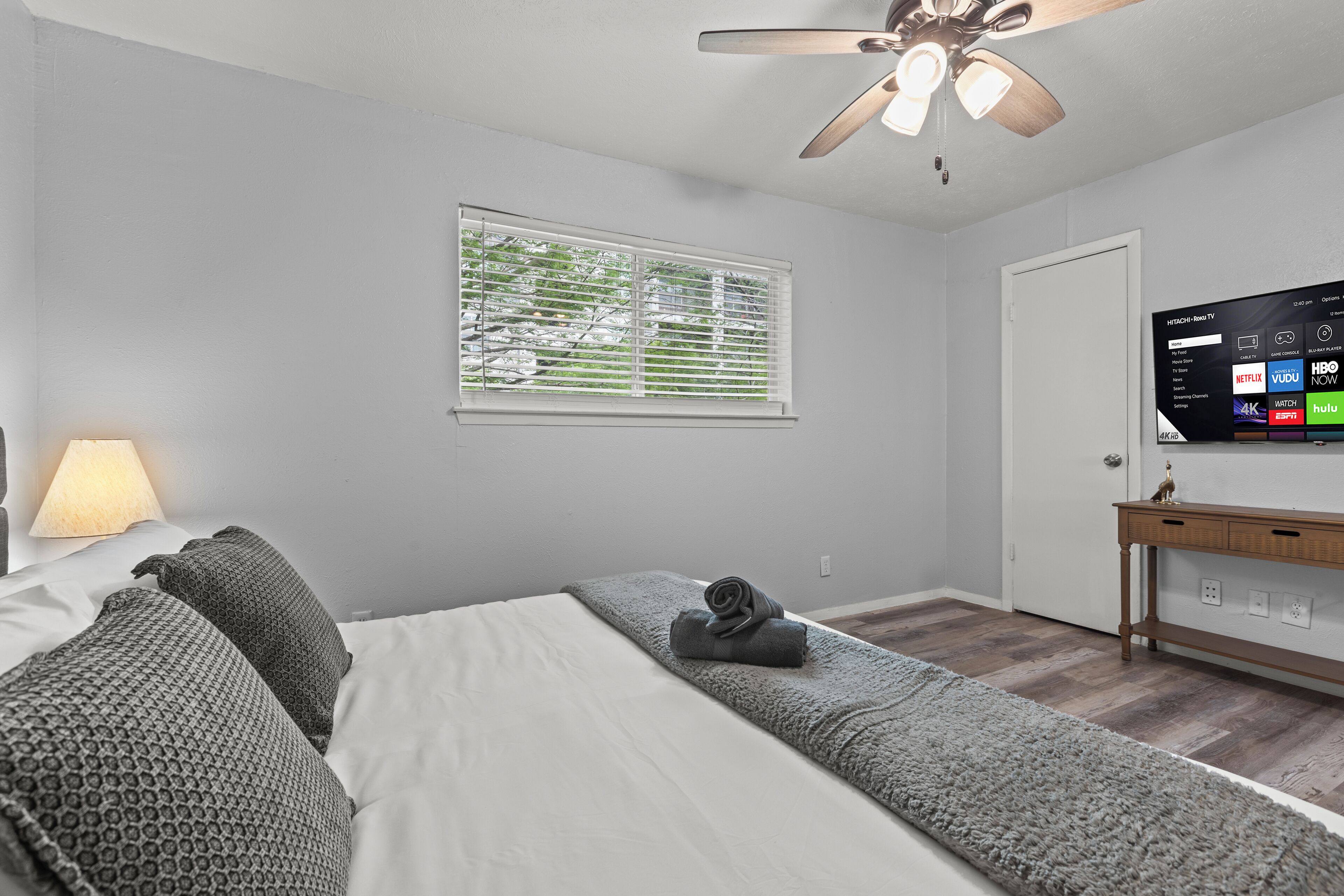Une chambre à coucher comprenant un lit, un téléviseur fixé au mur, un ventilateur de plafond, une fenêtre avec des stores et une lampe de chevet.