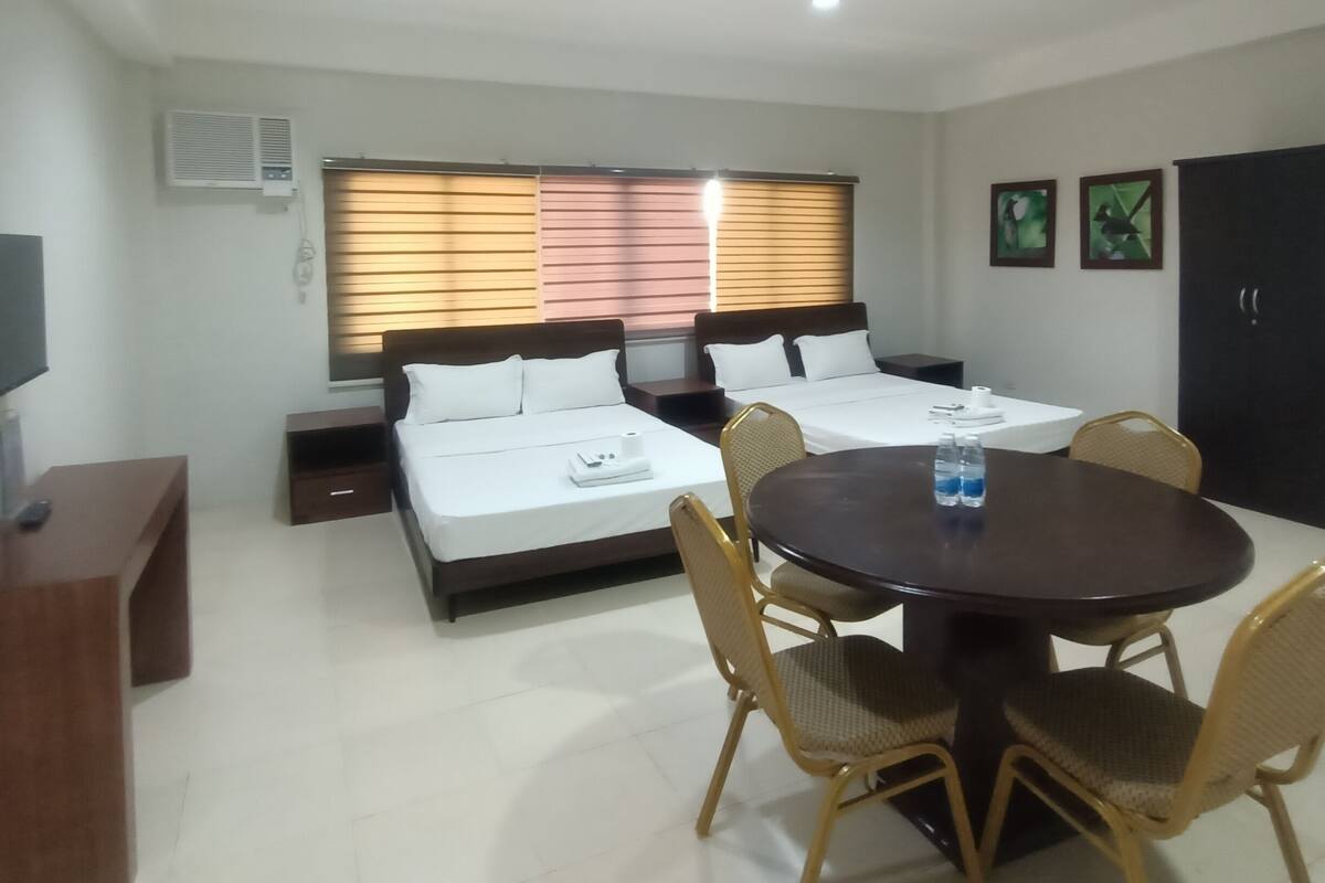 A hotel room with two beds, a dining table with chairs, a TV, and an air conditioning unit.