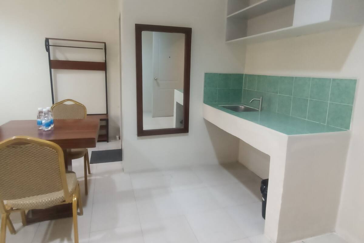 A small, clean room with a dining table, chairs, a mirror, and a kitchenette with a sink and shelves.